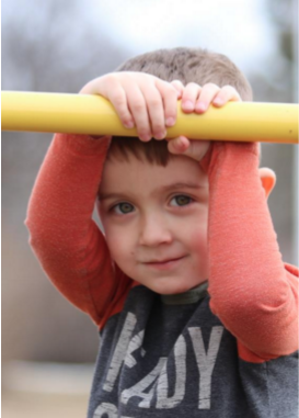 Photo of a little kid holding onto a yellow bar, arms framing the face. Photo by Melanie Young.