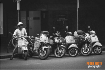 Photo of a row of scooters on the street with a gentleman sitting on one of them. Black and white. Street photography style.