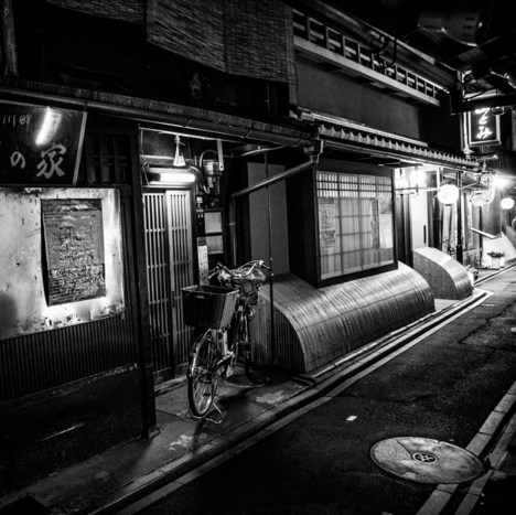 black and white photo of a bicycle parked outside a building in a street in old town kyoto