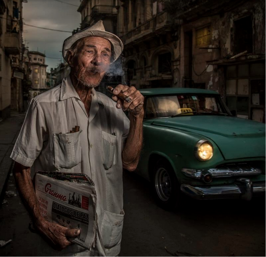 cuban man wearing a hat and shirt in front of a car and smoking