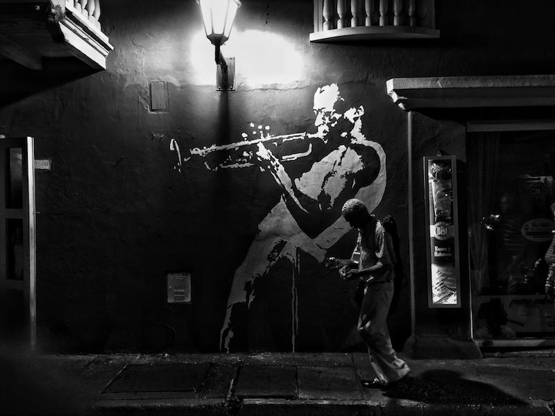Black and white photo. Man walks in front of a mural at night.