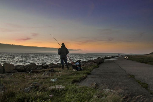 person fishing near the rocks with sunset over the water but slightly more orange