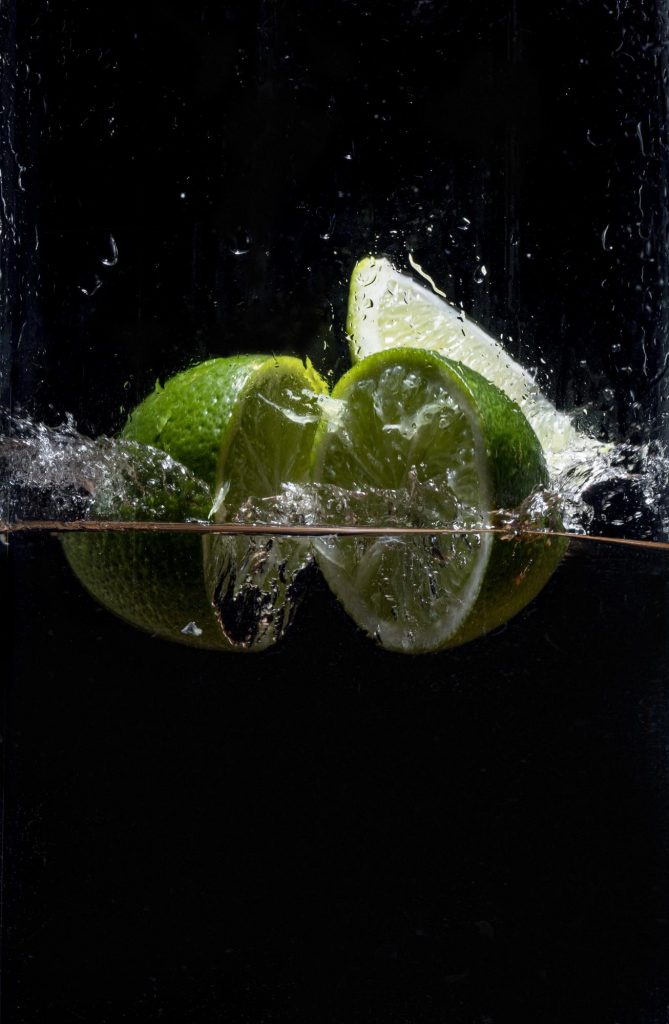 Two halves of a lime are breaking the surface of the water against a black background. You can see a wedge of lime in the background.