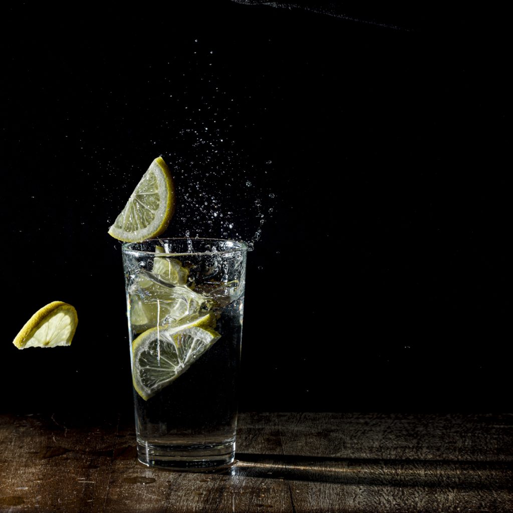 A glass of water against a black background with lemon slices being dropped into it, creating a splash. You can see droplets of water.