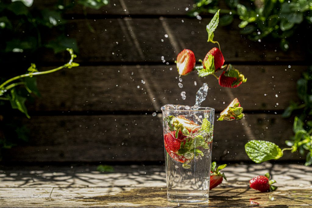 A glass of water with strawberries dropping into it creating a splash. This is taken in a garden with a wooden wall in the background and green vines. There is clear daylight filling the image. 